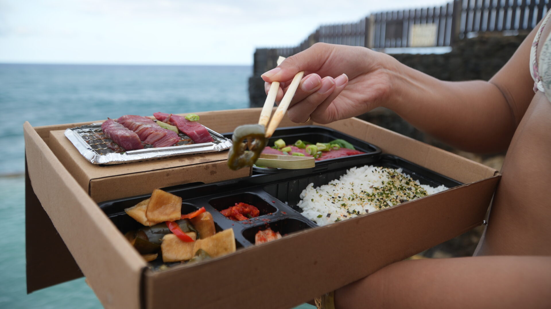 A person holding chopsticks over food in a box.