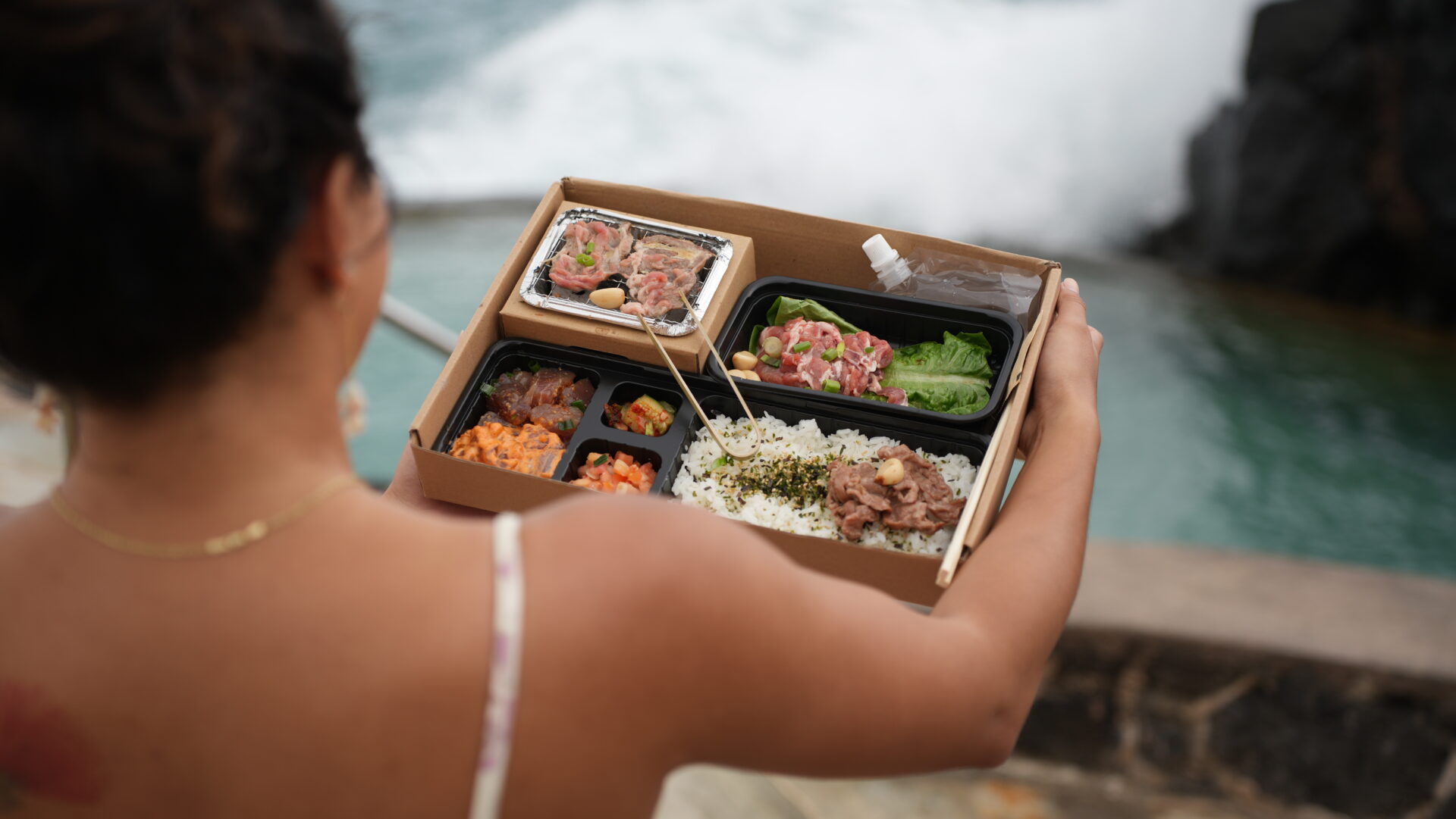 A woman holding a box of food in front of the ocean.